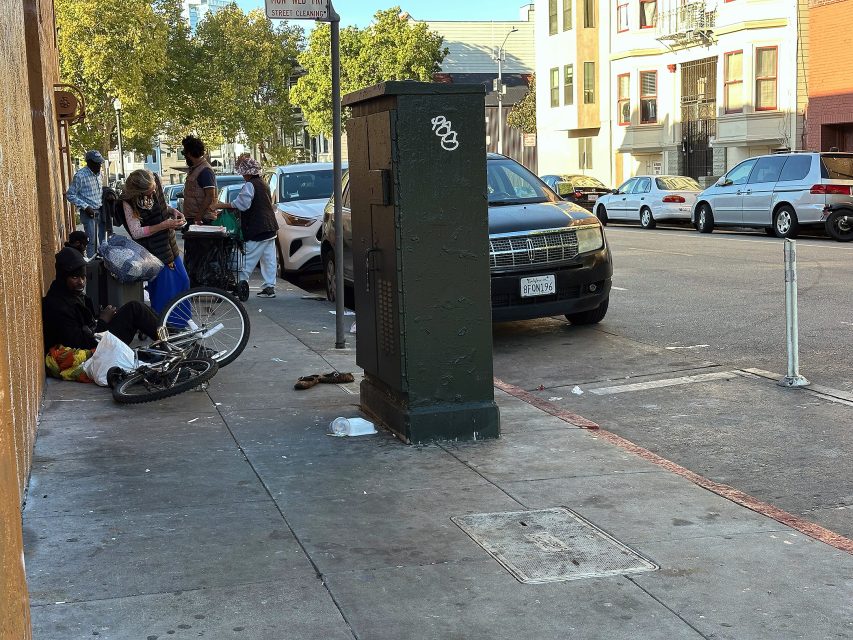 People gather on a city sidewalk near a utility box and a bicycle, with parked cars and apartment buildings visible in the background.
