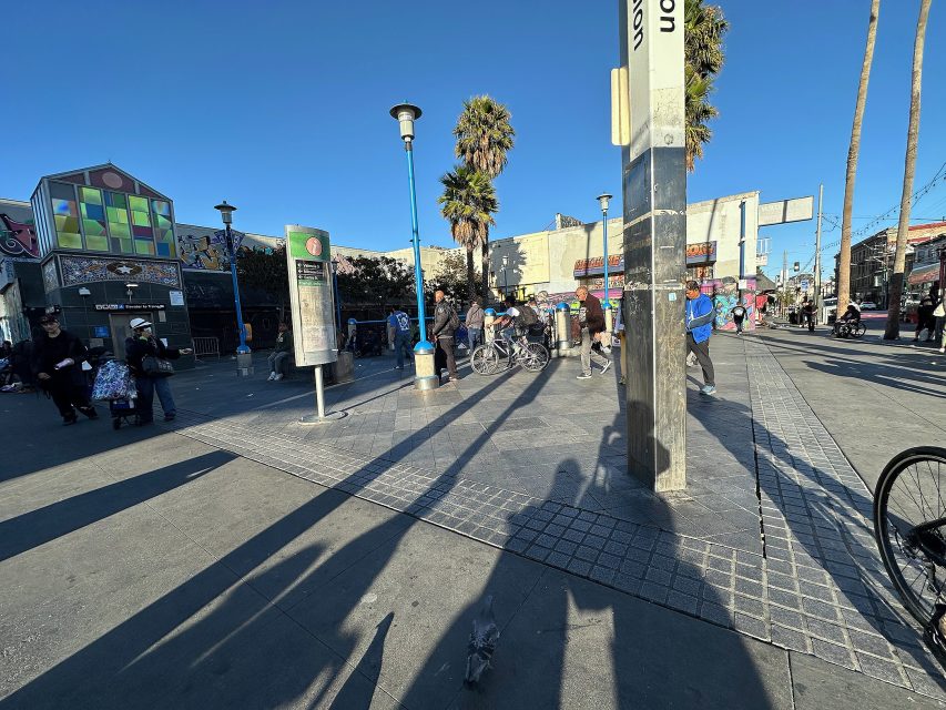 A busy outdoor plaza with people walking, biking, and sitting near palm trees and colorful buildings under a clear blue sky.