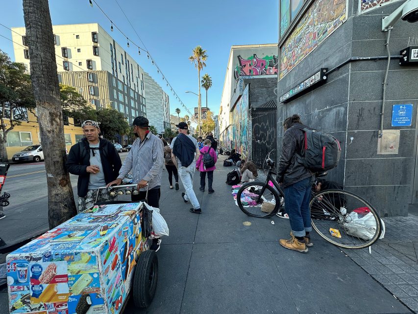 A city sidewalk with pedestrians, street vendors, a person repairing a bicycle, and people sitting near bags and belongings against a building wall.