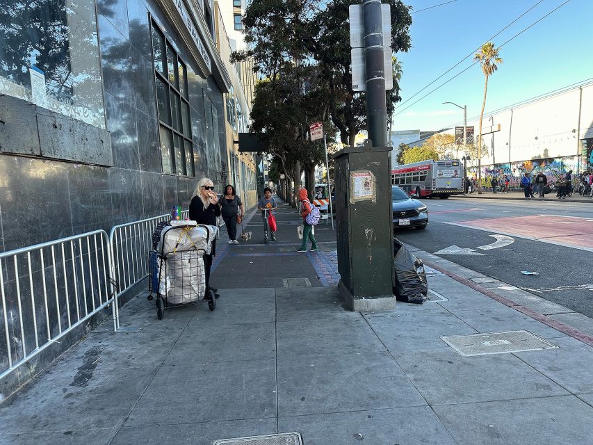 Several people stand and walk on a city sidewalk near a busy street, with a stroller, trash bag, and bus visible in the background.