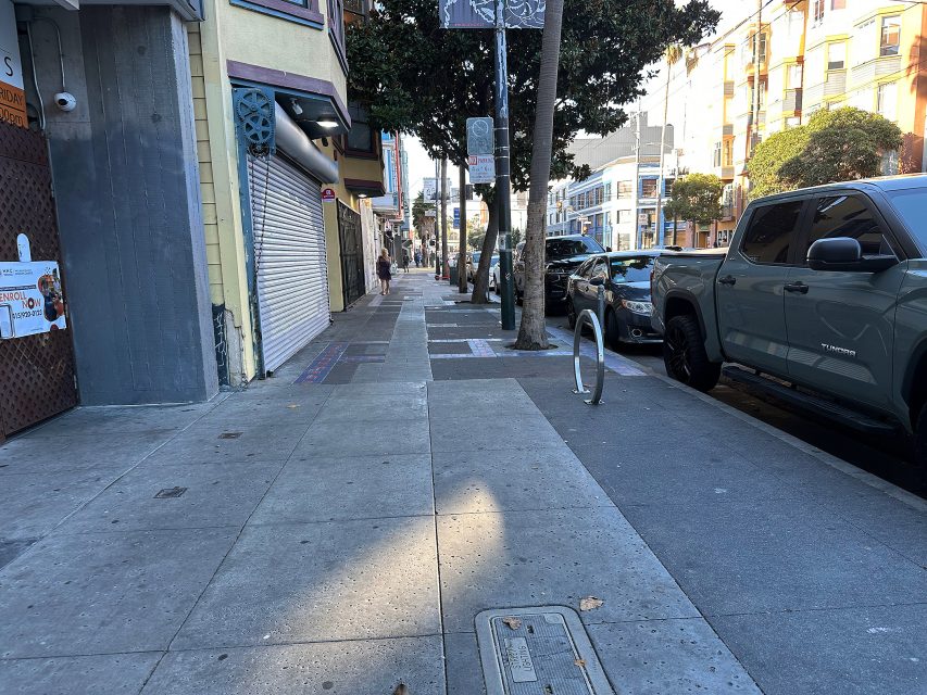 A city sidewalk with parked cars on the right, closed storefronts on the left, and a few people walking in the distance under daylight.