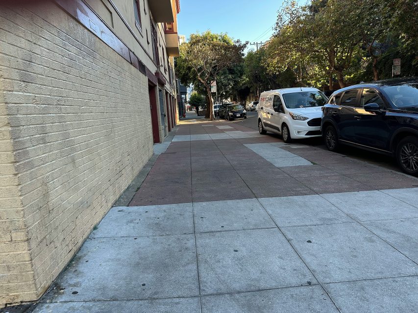 A city sidewalk runs alongside a beige brick building, with parked cars on the right and trees lining the street in the background under clear skies.