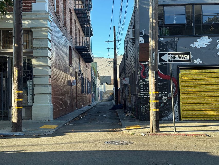 A narrow urban alleyway between buildings with murals, utility poles and a “One Way” street sign under clear daylight.