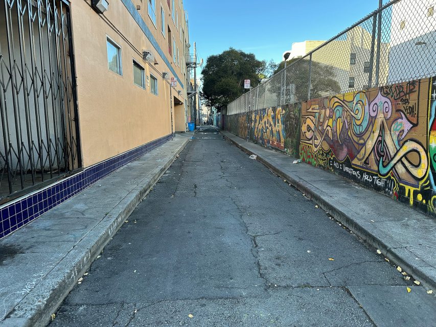 A narrow urban alley with cracked pavement, a beige building on the left, and a chain-link fence with colorful graffiti on the right, under a clear blue sky.