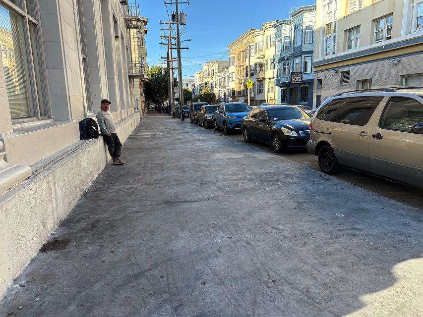 A man sits on a ledge along a wide sidewalk next to parked cars and multi-story buildings on a sunny day.