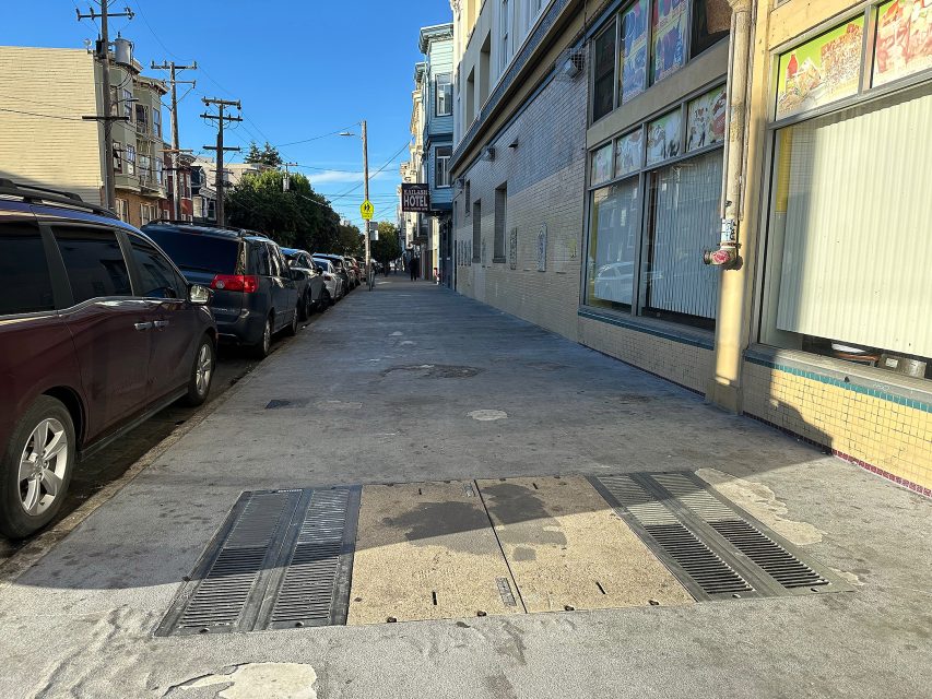 A city sidewalk beside parked cars and buildings, with a metal grate covering part of the pavement in the foreground. The sky is clear and it is sunny.