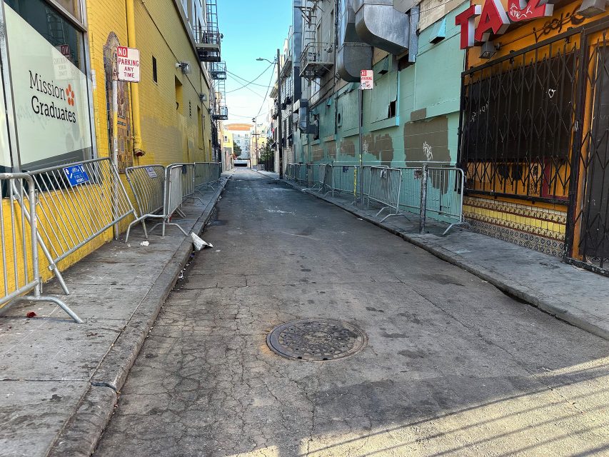 A narrow urban alleyway with metal barricades on both sides, cracked pavement, and buildings painted yellow and green.