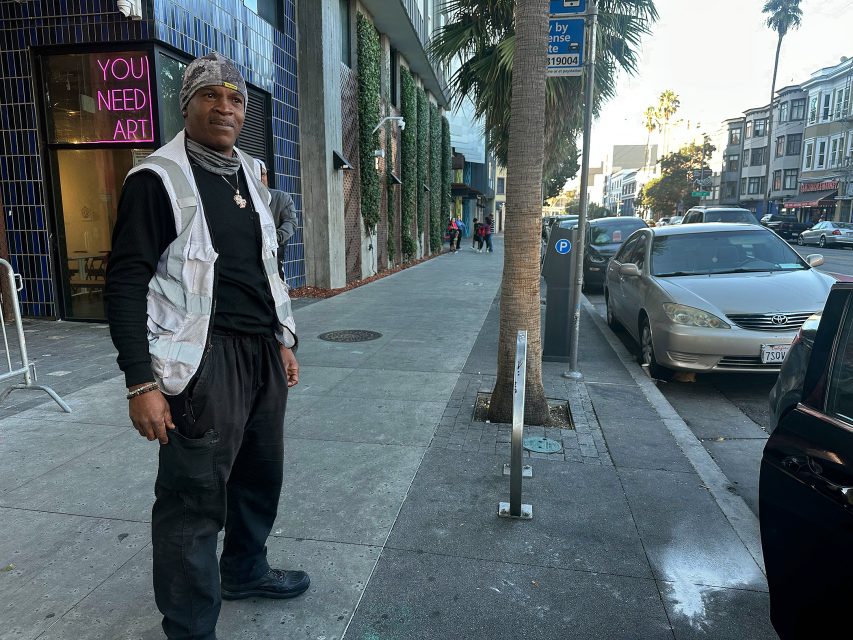 A man stands on a city sidewalk near a parking meter, with parked cars, palm trees, and a "YOU NEED ART" neon sign in the background.