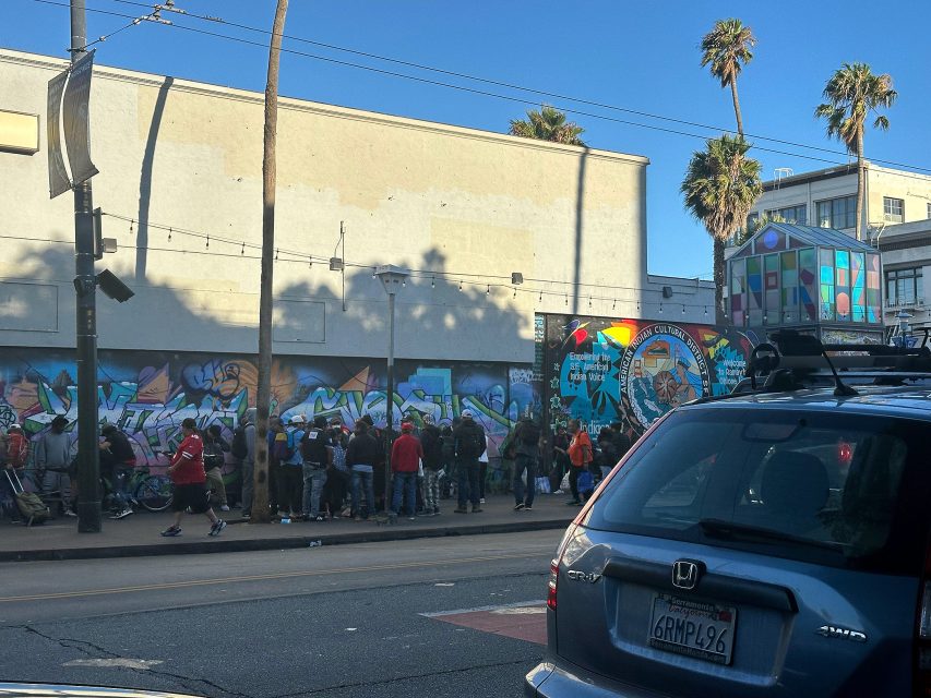 A group of people stand in line on a sidewalk beside a mural-covered wall in an urban area with palm trees; a dark Honda SUV is parked in the foreground.