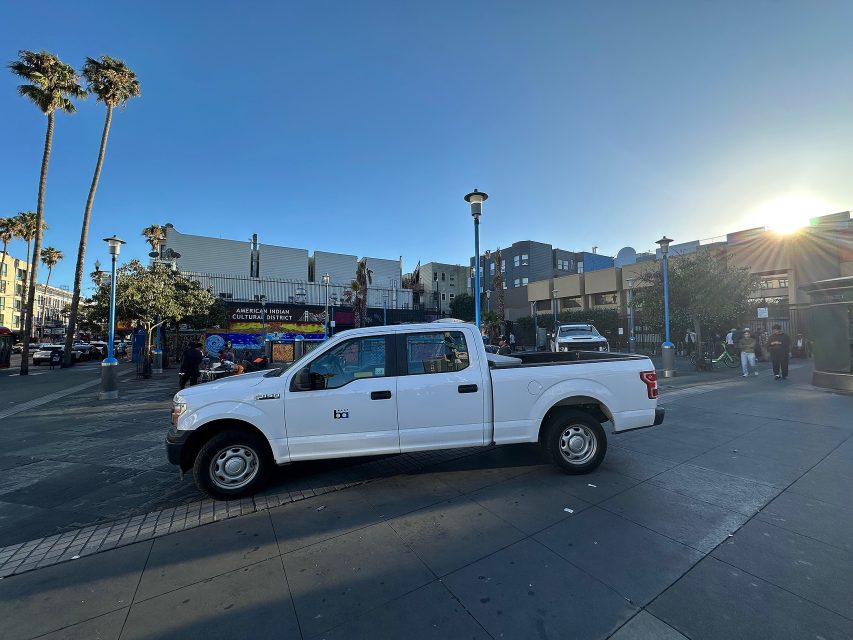 A white pickup truck is parked on a city plaza with palm trees, buildings, and pedestrians visible in the background under a clear blue sky.