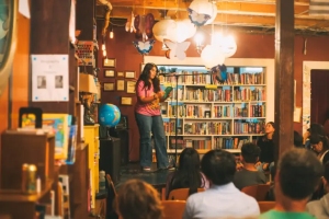 A person stands at a microphone reading from a book to an audience in a cozy, book-filled room with warm lighting and eclectic decor.