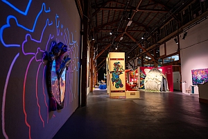 Art gallery interior with colorful wall installations, sculptures, and paintings displayed under ceiling lights in a spacious, industrial-style room.