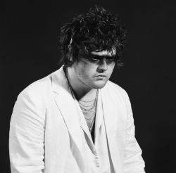 A person with dark, curly hair wears a white blazer, layered necklaces, and dark eye makeup, posing against a dark background.