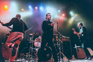 A four-member band performs energetically on stage with colorful lights in the background.
