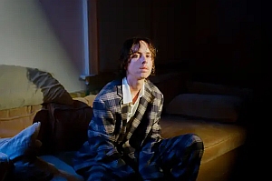 A person wearing a plaid suit sits on a couch in a dimly lit room, looking directly at the camera.