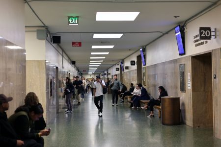 People stand and sit in a busy hallway of a public building with beige walls, overhead lighting, electronic screens, and EXIT signs visible.