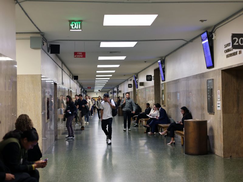 People stand and sit in a busy hallway of a public building with beige walls, overhead lighting, electronic screens, and EXIT signs visible.