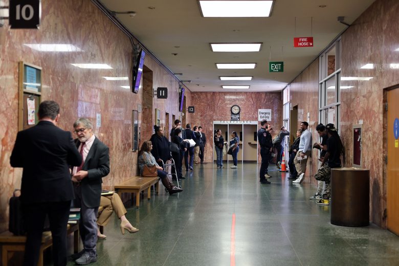 People wait and converse in a courthouse hallway with benches along the walls, officers present, and rooms numbered 10 to 13 visible.