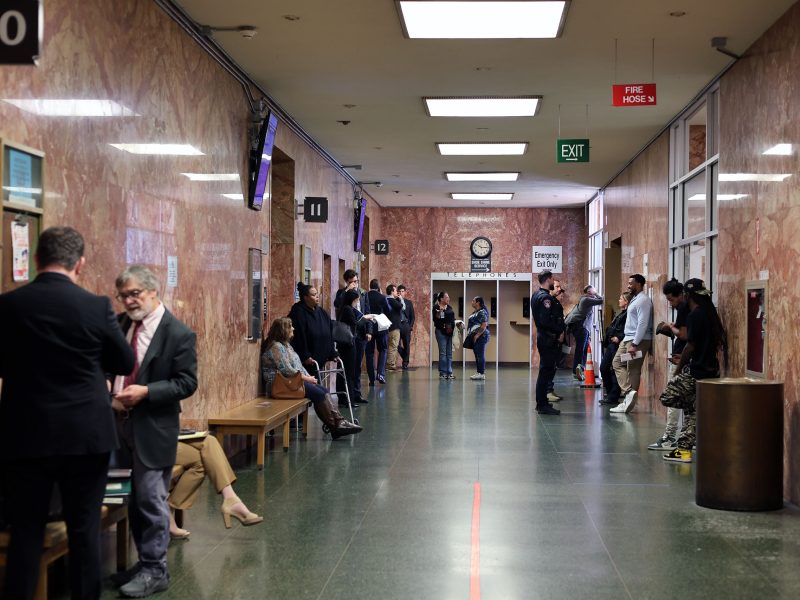 People wait and converse in a courthouse hallway with benches along the walls, officers present, and rooms numbered 10 to 13 visible.