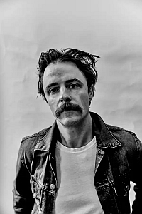 Black and white photo of a man with a mustache, tousled hair, wearing a denim jacket over a white shirt, looking directly at the camera against a plain background.