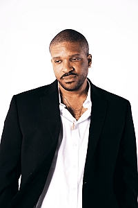 A man wearing a black blazer over a white shirt stands against a plain white background, looking slightly downward.