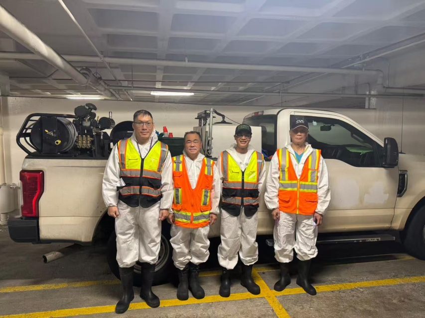 Four workers in white protective suits and reflective vests stand in front of a utility truck inside a parking garage.