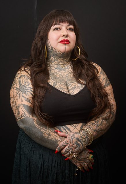 A woman with long brown hair, extensive tattoos, and gold jewelry stands against a black background, wearing a black top and skirt.