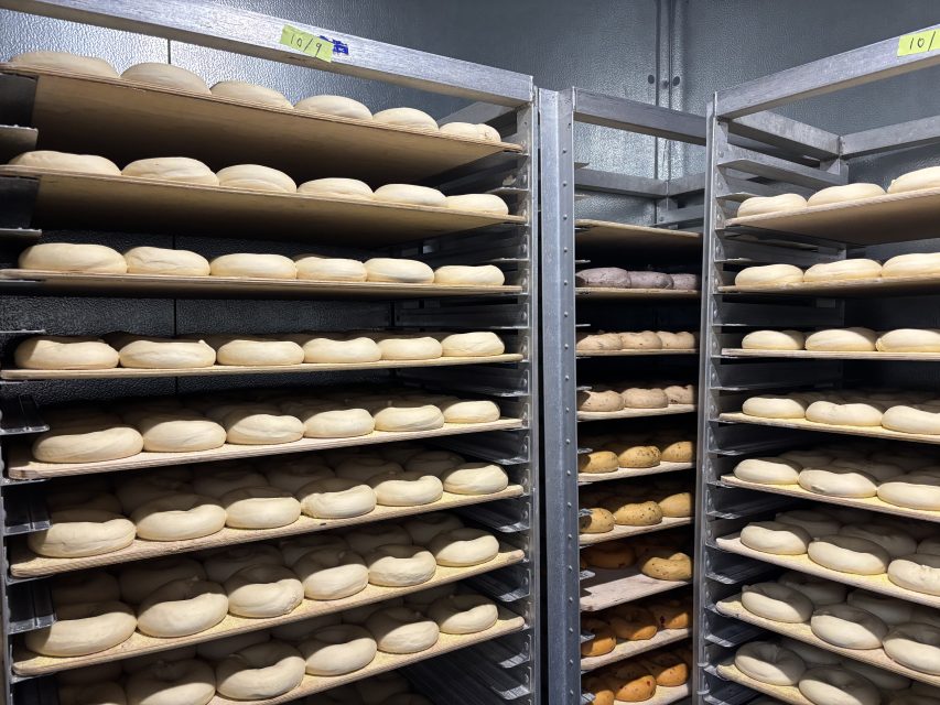 Multiple metal racks hold trays filled with rows of unbaked bread dough in a commercial bakery setting.