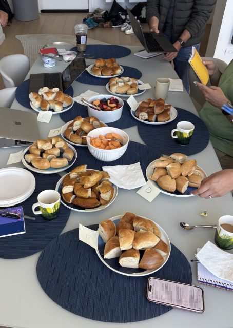 A table with several plates of assorted pastries and bread rolls, bowls of fruit, coffee mugs, laptops, and notebooks, with people standing nearby.