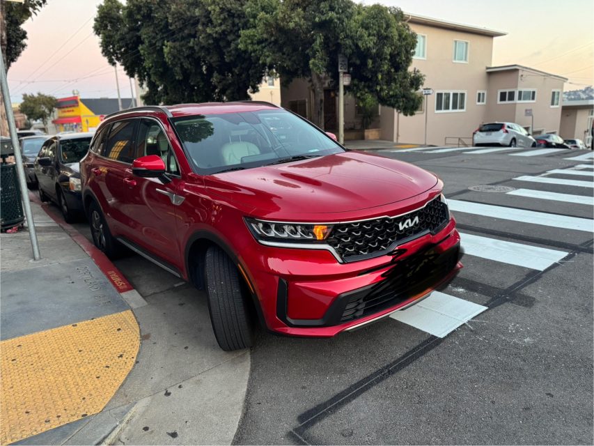 A red Kia SUV is parked partially on the sidewalk and crosswalk at a street corner near a residential area.