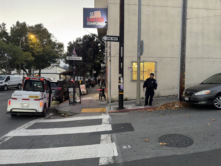 A police officer stands on a street corner near a crosswalk and a United Dumplings restaurant, with a parking enforcement vehicle parked nearby.