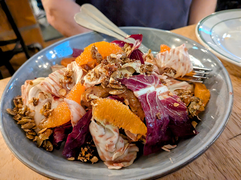 A salad with orange slices, radicchio, seeds, and a creamy dressing is served in a gray bowl with a fork and spoon.