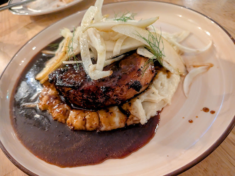 Grilled meat served on mashed potatoes with dark sauce, topped with thinly sliced vegetables on a beige plate.