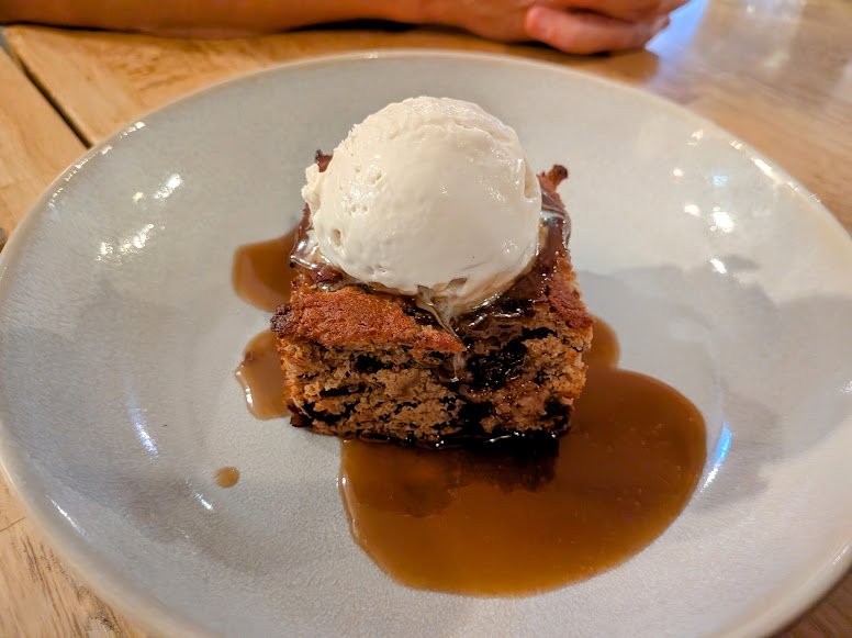 A scoop of vanilla ice cream sits on a slice of cake with brown sauce on a white plate, on a wooden table.