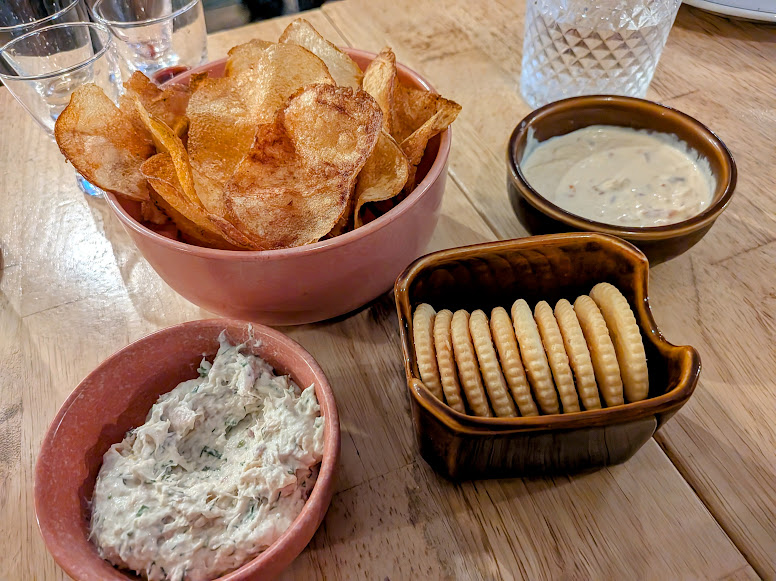 Bowls of potato chips, crackers, creamy dip, and a spread are arranged on a wooden table.