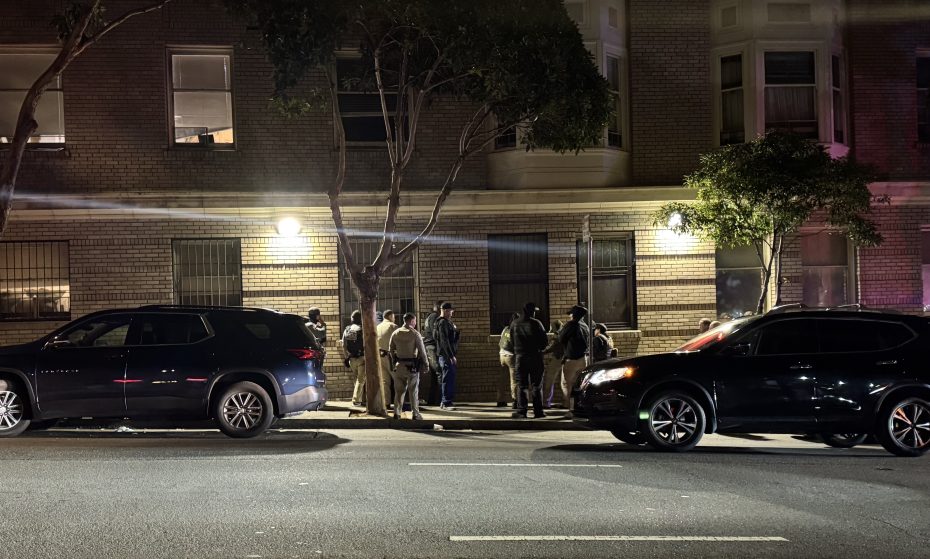 A group of police officers and people stand on a sidewalk at night in front of an apartment building, with two parked SUVs nearby.