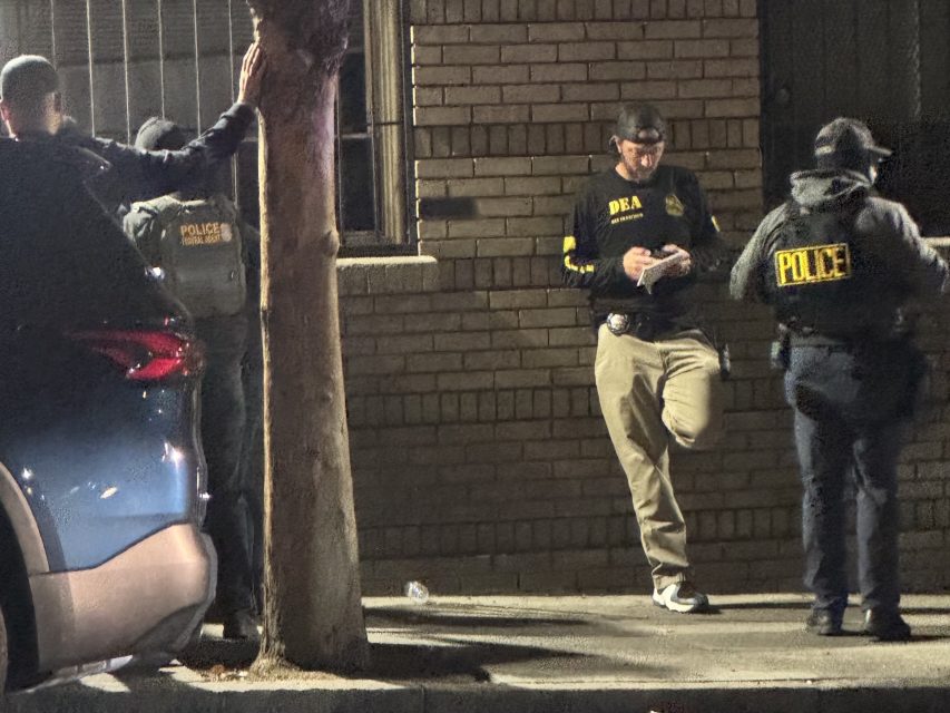 Three law enforcement officers stand on a sidewalk at night; two wear jackets labeled "POLICE" and one wears a jacket labeled "DEA.