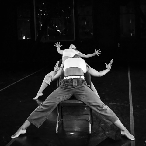 Three people sit on chairs in a dark room, leaning backward with arms outstretched, creating dynamic, intersecting lines with their bodies.