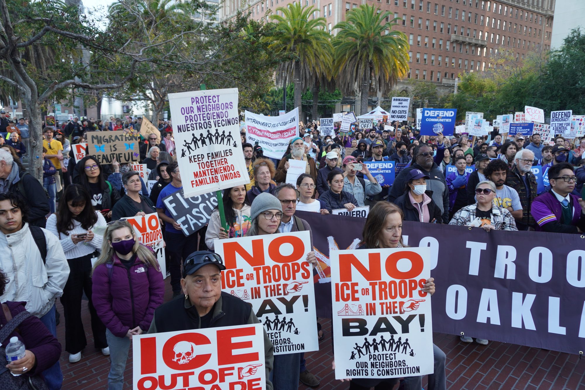 A large group of people hold signs opposing ICE and military presence in the Bay Area during a protest in an urban plaza, with palm trees and buildings in the background.