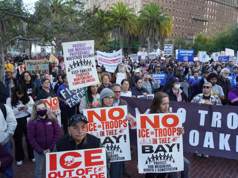 A large group of people hold signs opposing ICE and military presence in the Bay Area during a protest in an urban plaza, with palm trees and buildings in the background.