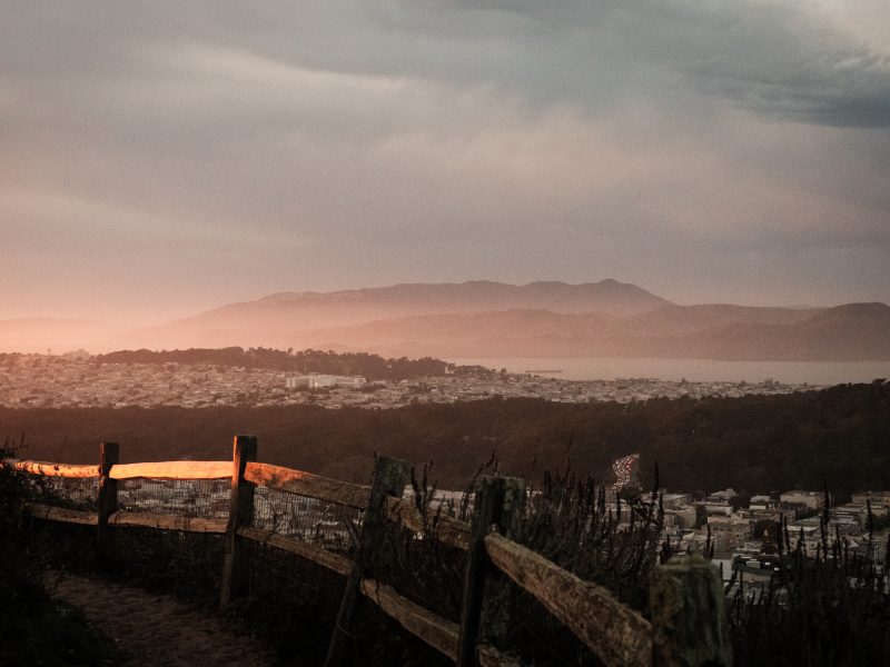 A dirt path with a wooden fence overlooks a city, forest, and distant mountains under a cloudy sky at sunset.