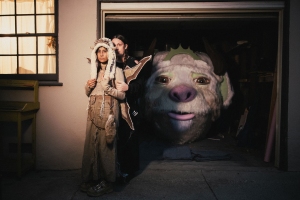 Two people in costumes stand in a garage doorway, with a large, animated creature's face visible behind them inside the garage.