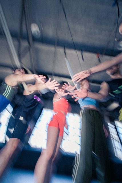 A group of people in athletic wear stand in a circle indoors, reaching their hands toward the center, viewed from below with a motion blur effect.