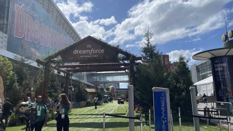 Entrance to Dreamforce National Park event area, with attendees walking on grass among trees and Salesforce branding under a sunny sky, capturing the Benioff vision for a connected community.