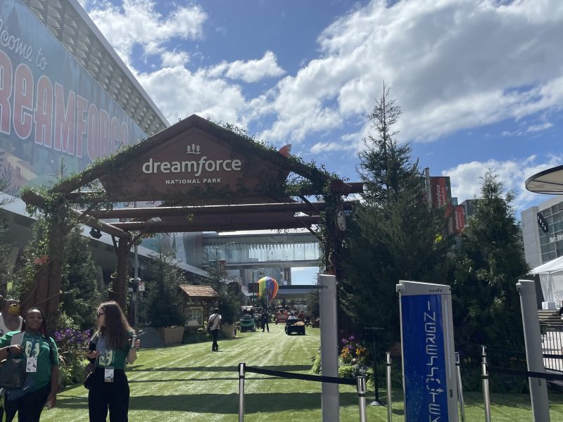 Entrance to Dreamforce National Park event area, with attendees walking on grass among trees and Salesforce branding under a sunny sky, capturing the Benioff vision for a connected community.