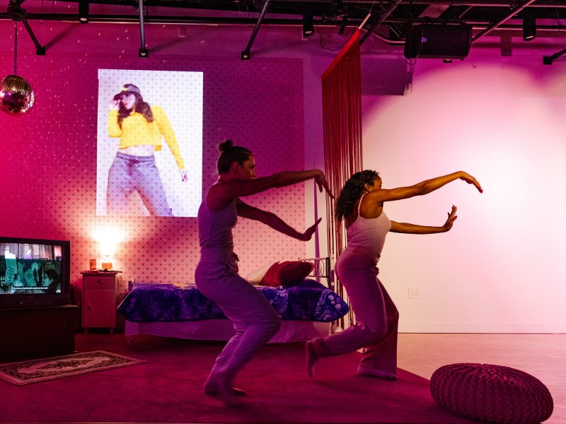 Two people dance in a room with a bed, TV, and projected video of a performer in yellow. The lighting is pink and purple, creating a lively atmosphere.
