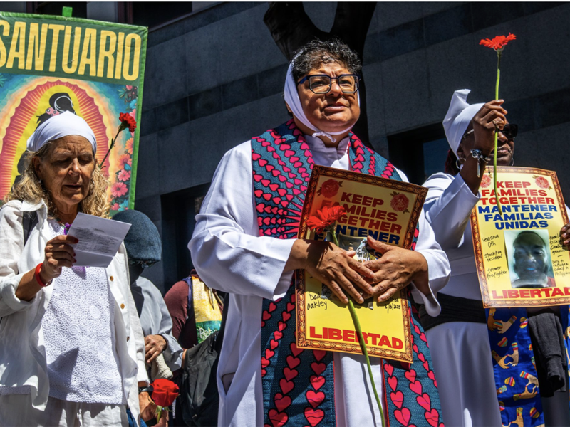 A group of women at a protest hold signs reading "Keep Families Together" and "Libertad," with one holding a red carnation. A "Santuario" banner is visible in the background.