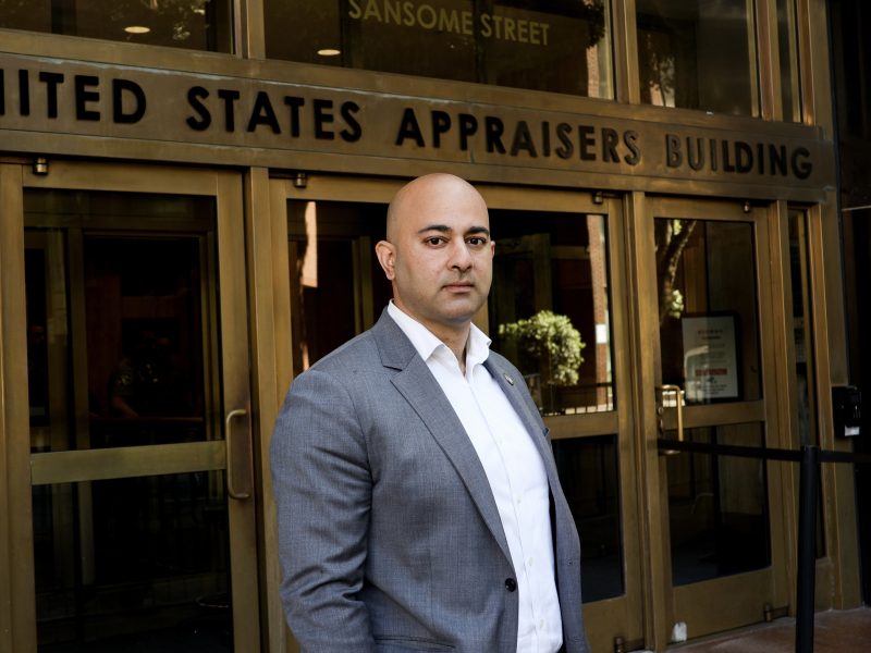 A man in a gray suit stands in front of the ICE entrance at the United States Appraisers Building.