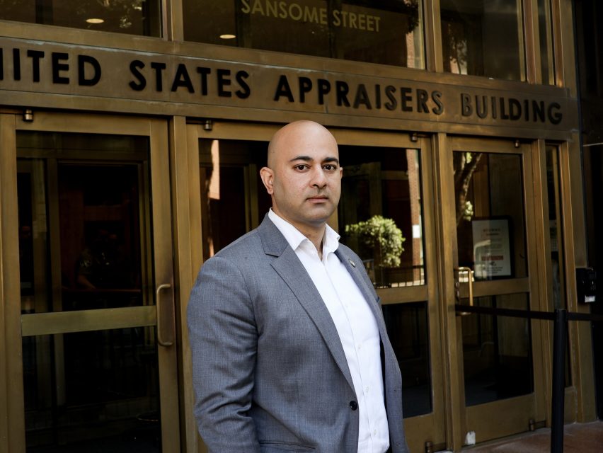 A man in a gray suit stands in front of the ICE entrance at the United States Appraisers Building.