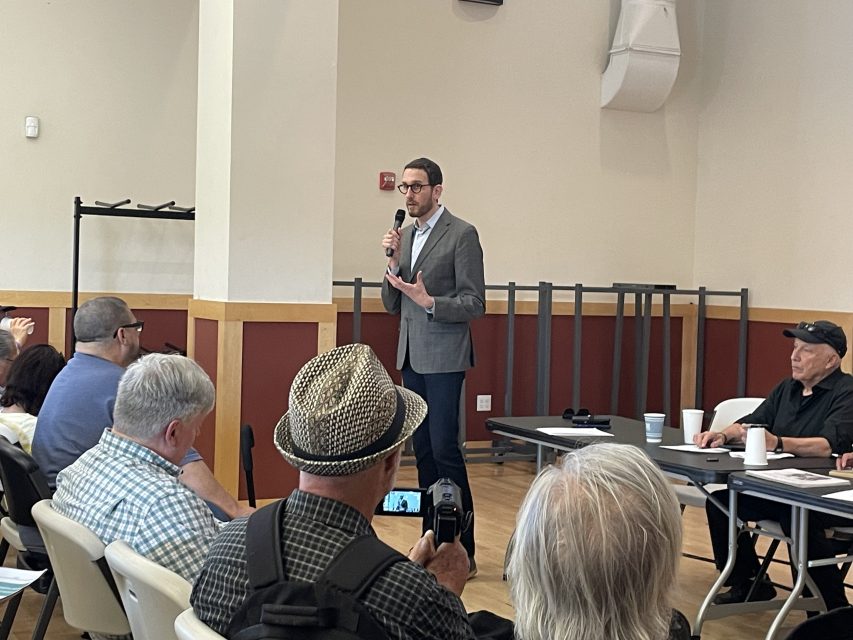 A man stands and speaks into a microphone in front of seated people at a community meeting in a room with red and white walls.